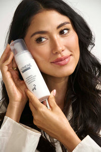 Beautiful woman with clear skin holding L'ATHENE Citrus Foam Exfoliating Cleanser bottle, smiling naturally.